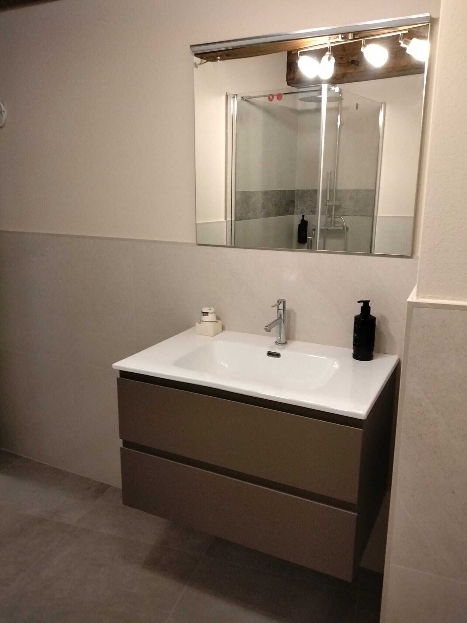 Une salle de bain avec un lavabo blanc, un miroir et une douche que l’on aperçoit dans le reflet.