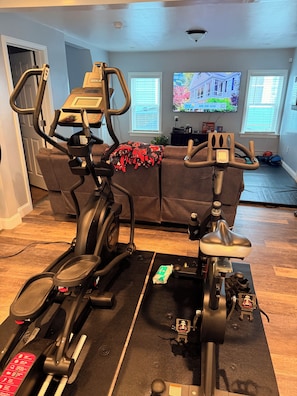 Ein Fitnessbereich im Innenbereich mit zwei Ergometer, einer Couch und einem Fernseher im Hintergrund.