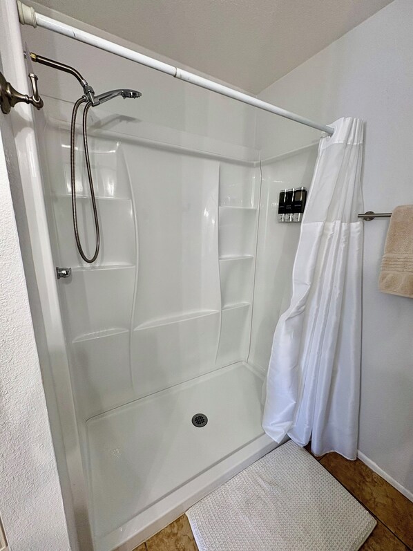 A glass-enclosed shower with a handheld showerhead, a white shower curtain, and a small shelf with toiletries. - Cozy 2-bedroom Golf course get away close to hospitals and attractions (Glendale)