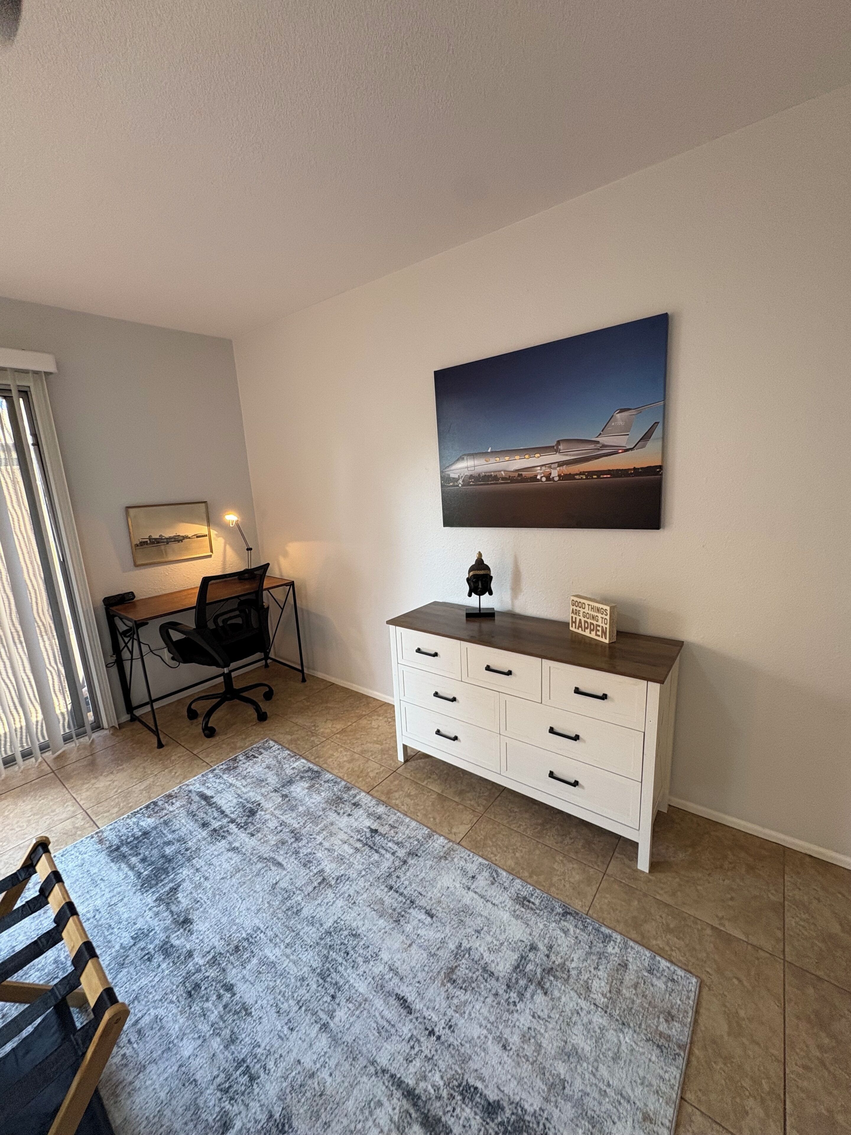Een kamer met een bureau, een stoel, een witte dressoir, een vloerkleed en een schilderij aan de muur.