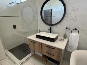 A modern bathroom with a rectangular sink, wooden cabinetry, a glass shower enclosure, and a round mirror.