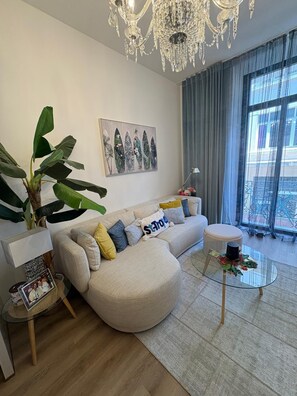 Ein modernes Wohnzimmer mit einer großen Pflanze, einem Sofa mit bunten Kissen, einem Glastisch und einer Kristallleuchter.