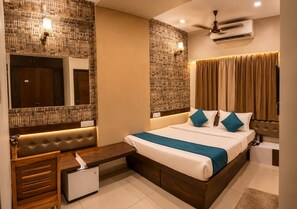 A hotel room with a large bed, a vanity with a mirror, a small desk, a ceiling fan, and an air conditioning unit. - Kubo By Convivial Hotels (Indore)