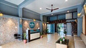 Zona de recepción de un hotel con mostrador, asientos, plantas en macetas y un ventilador de techo.