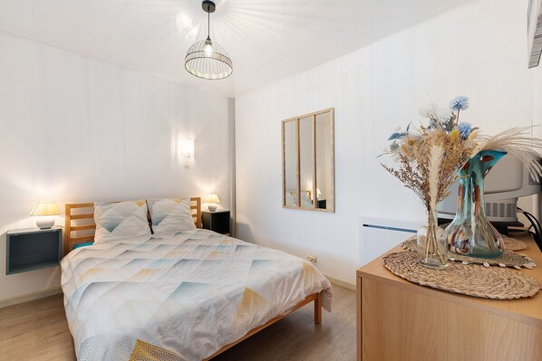 A bedroom with a bed, bedside tables, a mirror, a desk with a vase, and a hanging light fixture.