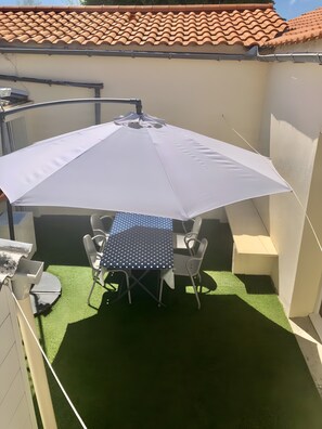 En uteplats med ett bord och stolar under ett vitt parasoll, omgiven av konstgjord, grön gräsmatta.