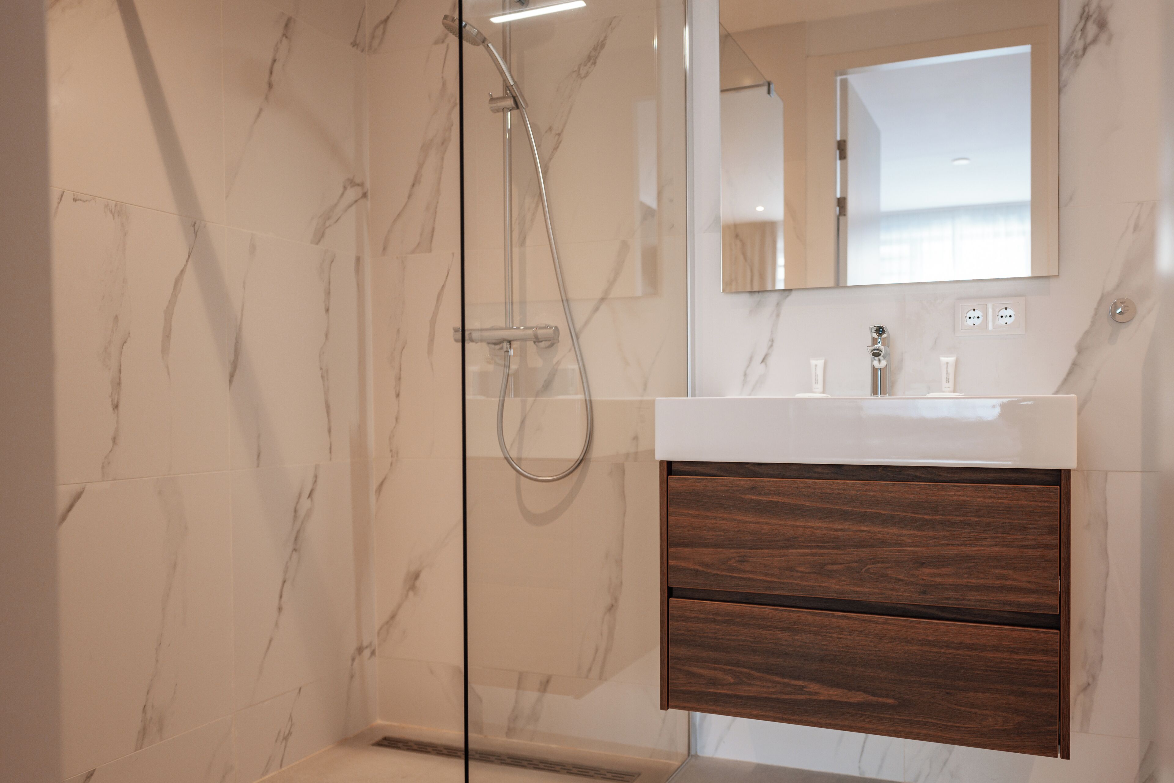 A modern bathroom with a glass-enclosed shower, marble walls, and a wooden vanity with a mirror above it.
