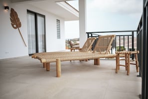 Ein Balkon mit einem gewobenen Liegestuhl, einem hölzernen Hocker und einer dekorativen Wandgestaltung mit Blättern.