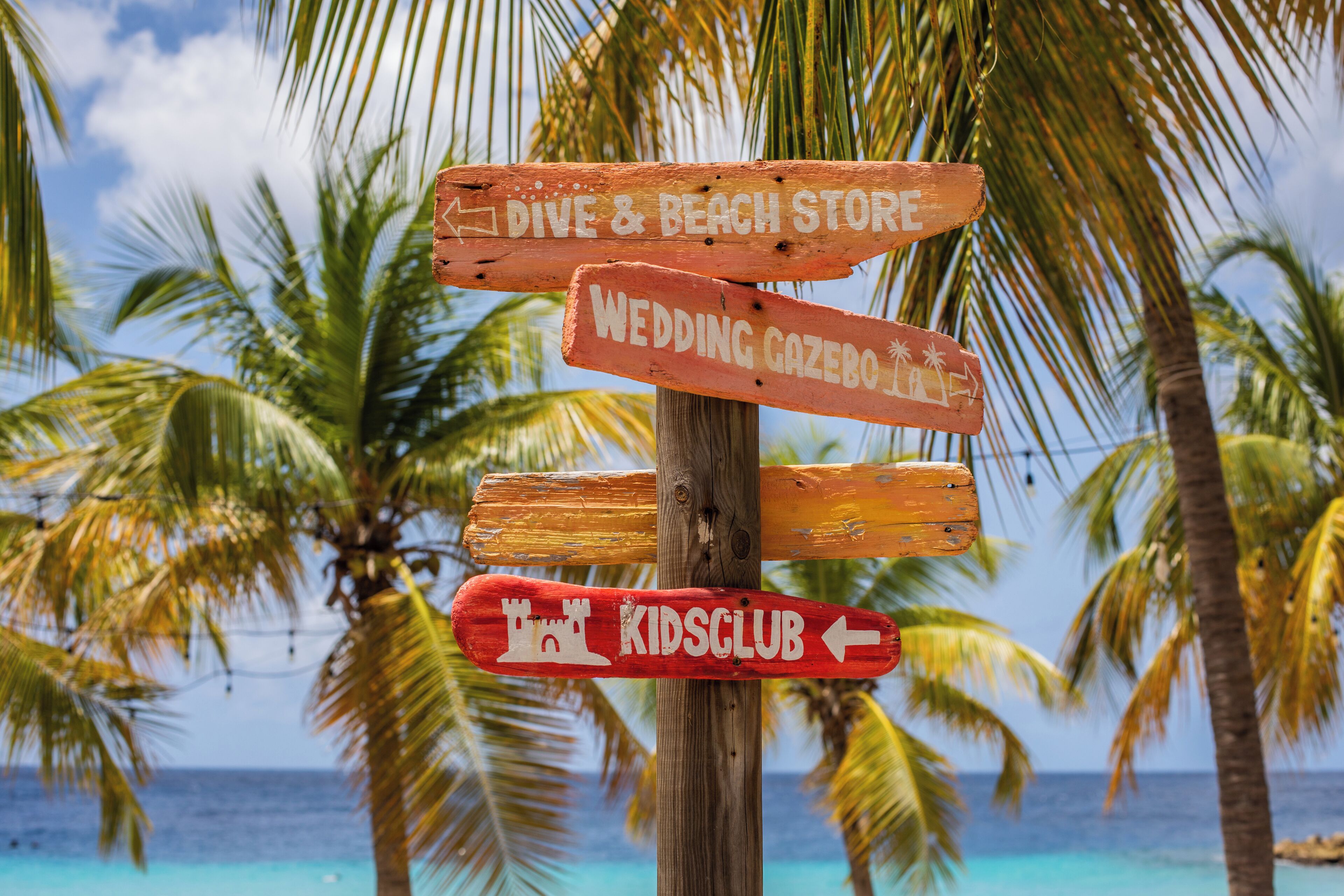 Un cartel de madera indica direcciones a una tienda de buceo y playa, un pabellón de bodas y un club infantil.