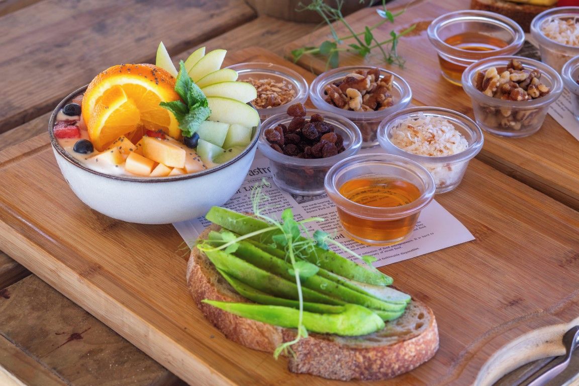 A wooden table with a bowl of fruit salad, a slice of bread with peas, and small containers of nuts and honey.