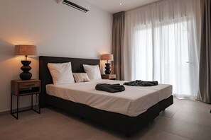 A modern hotel room with a large bed, two bedside tables with lamps, a window with curtains, and a wall-mounted air conditioner. - The Breeze -Blue Bay Golf & Beach Resort (Sint Michiel)