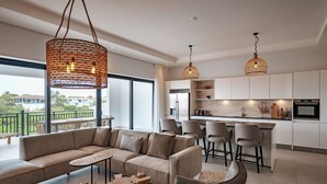 Ein modernes Wohnzimmer mit einer Couch, einem Couchtisch und Sesseln, eine Küche mit Barhocker und große Fenster mit Blick ins Freie.