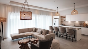 Ein modernes Wohnzimmer mit Sofa, Couchtisch und Sesseln, eine Küche mit Barhocker und Pendelleuchten.