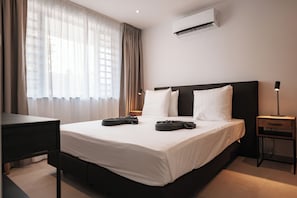 Ein Hotelzimmer mit Bett, Schreibtisch, Nachttisch, Klimaanlage und einem fenster mit Vorhängen.
