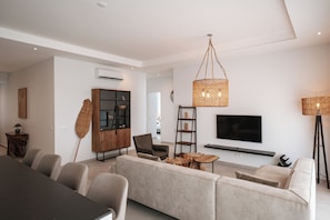 Un salon moderne comprenant un canapé, des fauteuils, une table à manger, un téléviseur et un grand luminaire décoratif en fibres tressées.
