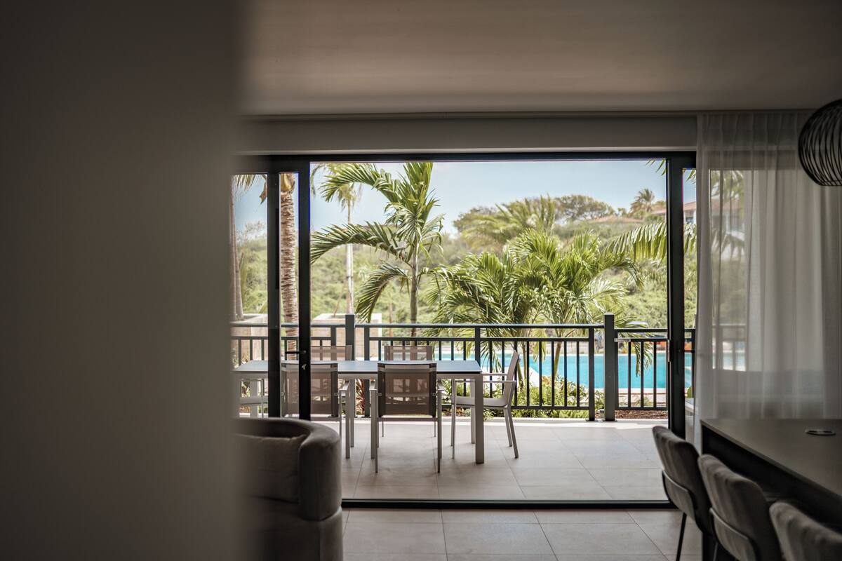 Una habitación con un gran ventanal corredero que da a un balcón con piscina y plantas tropicales.