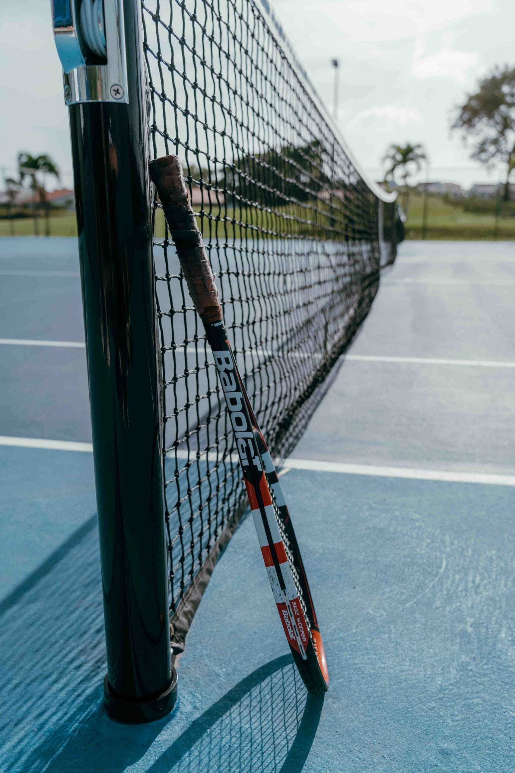 Una red de tenis con una raqueta apoyada, un poste metálico y una superficie de juego azul.
