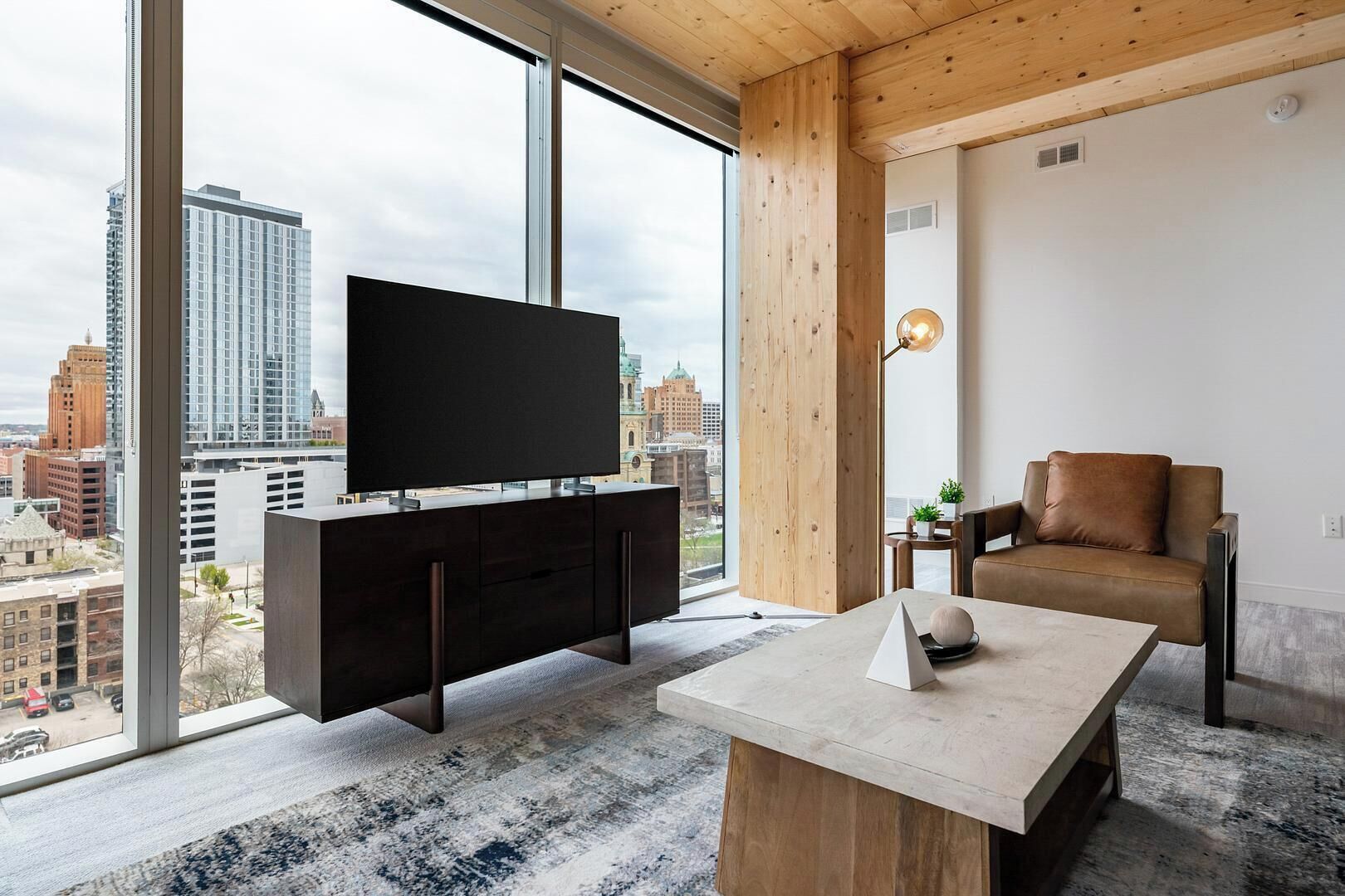 A modern living room with a large TV, a wooden coffee table, a leather sofa, and a city view through large windows.