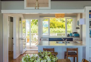 A kitchen with a large island, a dining area with two chairs, a view of the outdoors, and a white railing. - Barefoot Bach Absolute Waterfront in Arran Bay (Waiheke Island)
