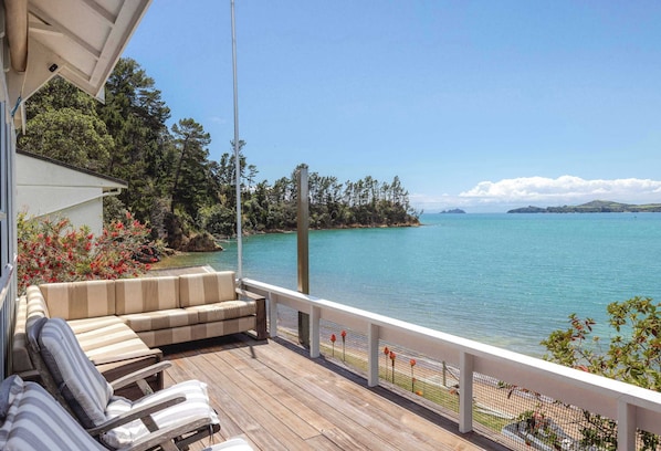 A wooden deck with a sofa and chairs, overlooking a calm lake with a forested island in the distance. - Barefoot Bach Absolute Waterfront in Arran Bay (Waiheke Island)