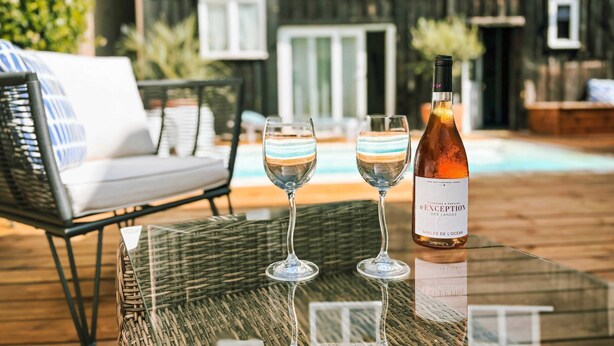 A glass table with two wine glasses and a bottle of wine, a wicker chair, and a pool in the background. - Villa Neptune: Family home in the city center (La Teste-de-Buch)
