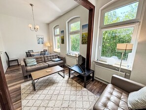 Ein Wohnzimmer mit Sofa, Couchtisch und Fernseher. Fenster bieten Blick auf Grünflächen.