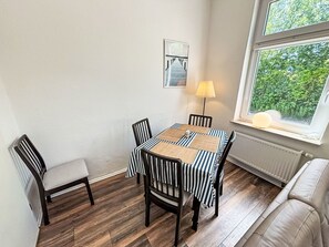 A dining area with a table and chairs, a lamp, a radiator, and a window with a view of greenery. - Wonderful  villa for 6 people with WIFI, TV, terrace and pets allowed (Norden)