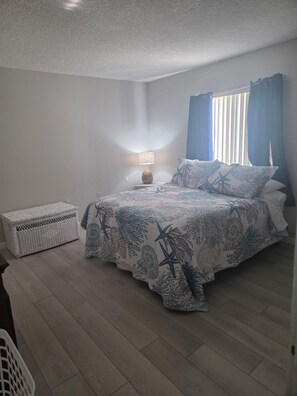 Ein Schlafzimmer mit einem Bett, einem Nachttisch, einer Lampe, einer weißen Korbo und einem Fenster mit Vorhängen.
