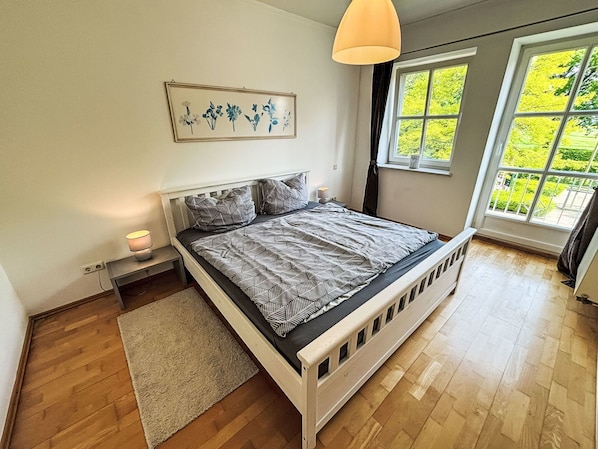 Un dormitorio con cama, mesitas de noche, alfombra, ventana con vistas a zonas verdes y un cuadro colgado en la pared.
