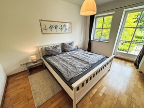 Ein Schlafzimmer mit einem Bett, Nachttischen, einem Teppich, einem Fenster mit Blick auf Grünflächen und einem Wandbild.