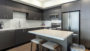 A modern kitchen with dark cabinets, a stainless steel refrigerator, a microwave, and a dishwasher. There is a light-colored island with two stools. - Landing Furnished Apartments Dallas along West Colorado Blvd (Dallas)