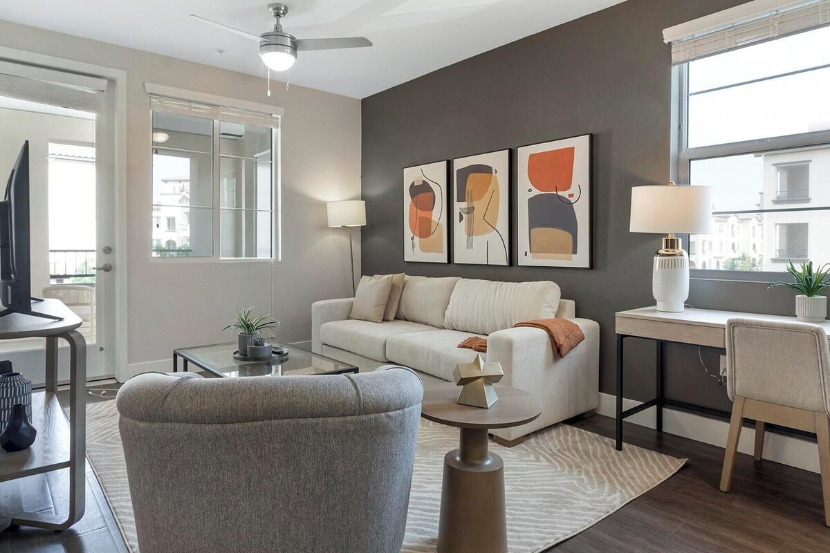 A modern living room with a sofa, coffee table, and a desk with a lamp. There are two wall-mounted artworks and a ceiling fan.
