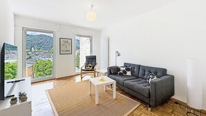 Amplia sala de estar con sofá, sillón, una mesita y televisor. Grandes ventanales ofrecen vistas a zonas verdes y edificios.