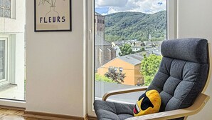 Una habitación con un ventanal amplio, una obra de arte enmarco con texto, una silla gris con cojín y vistas a un pueblo y montañas.