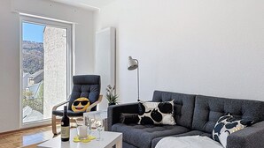 Un salón con un sofá gris, una silla, una mesa con una botella y vasos, una lámpara, y vistas a través de una puerta.