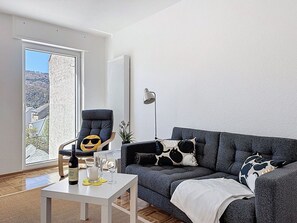 Ein Wohnzimmer mit grauem Sofa, einem Sessel, einem Tisch mit Flasche und Gläsern, einer Lampe und Blick durch eine Tür.