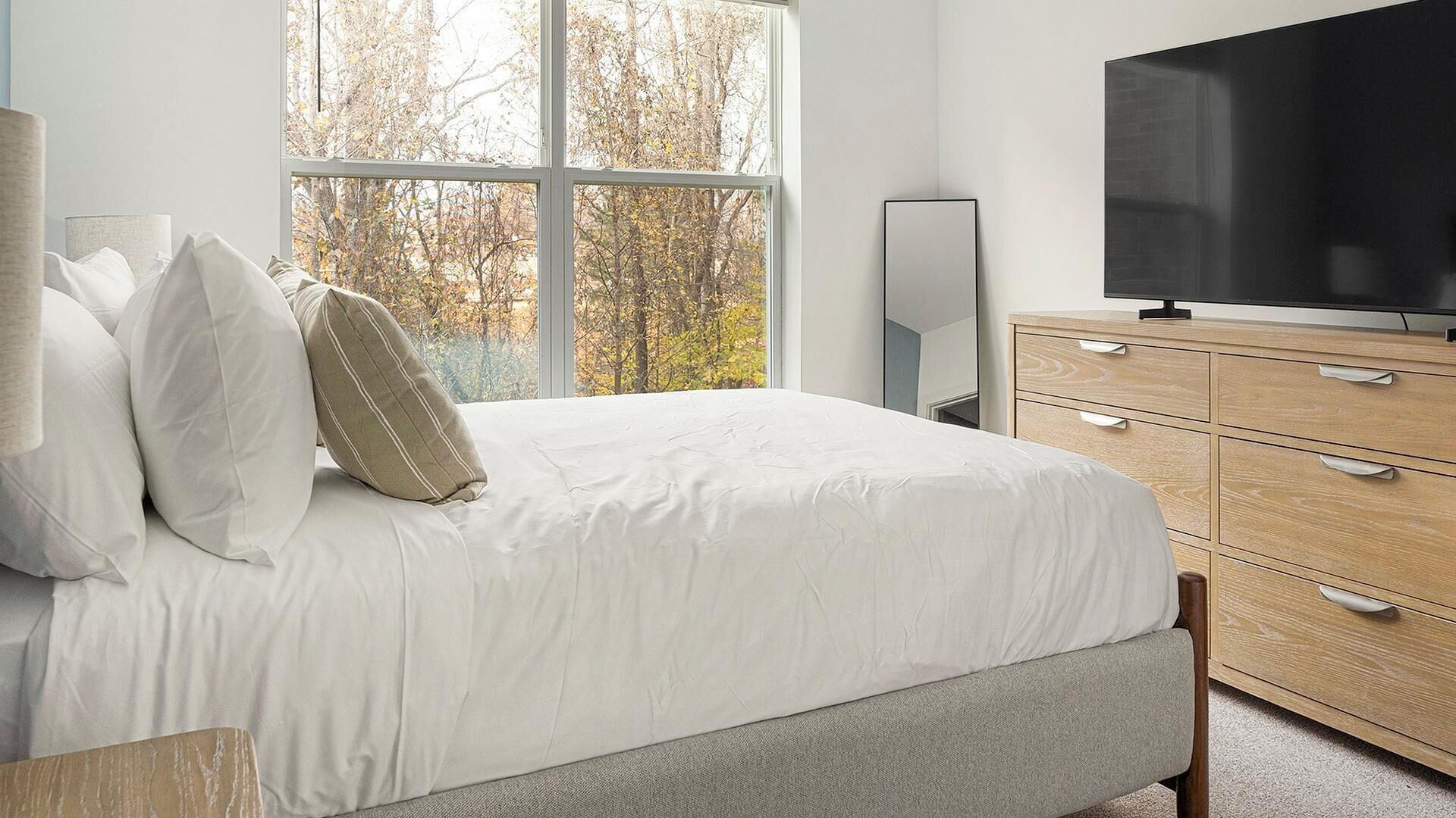 A bedroom with a large window, a bed with white bedding, a TV, and a wooden dresser.
