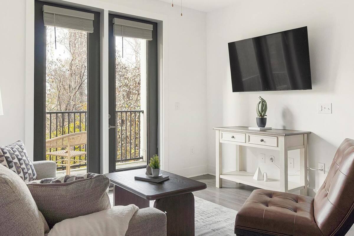 A modern living room with a sofa, a brown leather chair, a small table with a plant, a white console table, and a TV mounted on the wall.