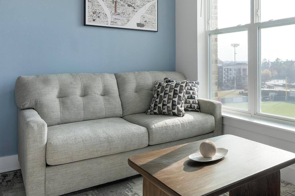 A living room with a grey sofa, patterned cushions, a wooden coffee table, and a city view through a large window.