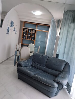 Ein Wohnzimmer mit einem schwarzen Ledersofa, einem Esstisch mit Stühlen, einem Bücherregal und einer Wand mit blauen Blumenmustern.