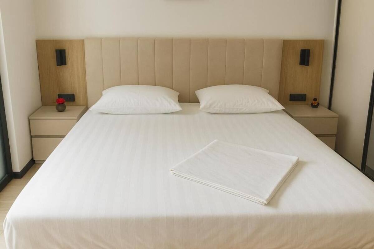 Una cama bien hecha con un cabecero color crema, dos mesitas de noche con adornos pequeños y una toalla blanca doblada sobre la cama.