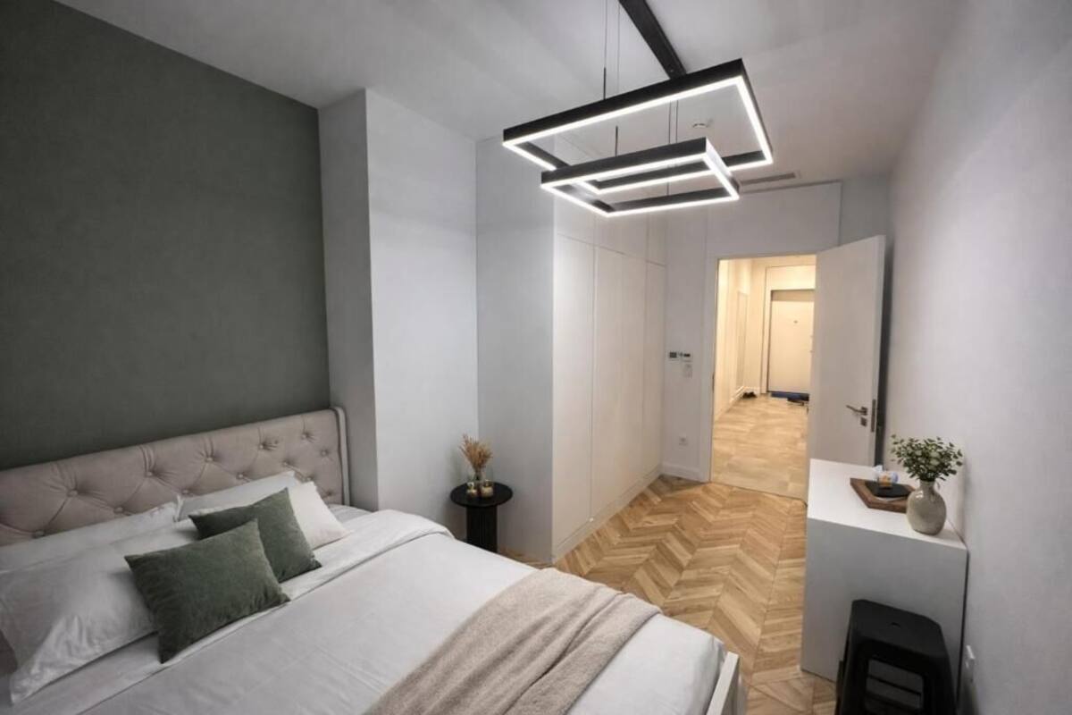 Un dormitorio moderno con cabecera capitonada, cama con ropa blanca y almohadas verdes, mesita de noche con un florero y suelo de madera con patrón de espiga.