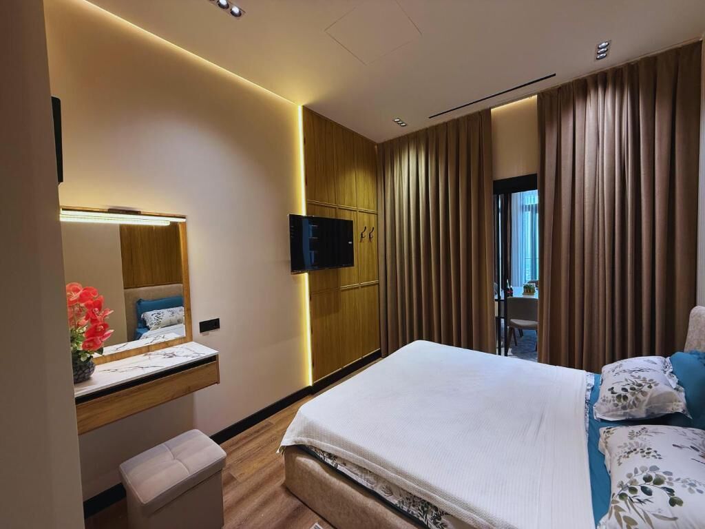 A modern hotel room with a bed, a desk with a mirror, a television, and a window with curtains.