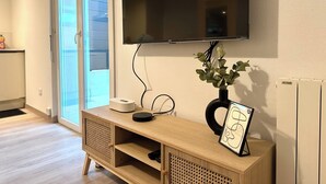 Ein modernes Wohnzimmer mit Flachbildfernseher an der Wand, einem Holztischchen mit Vase und Bilderrahmen sowie einer Glasschiebetür, die zu einem Außenbereich führt.