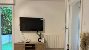 Ein modernes Wohnzimmer mit einem an der Wand montierten Flachbildfernseher, einer Holzunterhaltungsecke, einer Glastür zu einem grünen Außenbereich und einem Esstisch mit Weingläsern und einer Weinflasche.