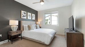 A bedroom with a bed, bedside lamps, a ceiling fan, a TV on a wooden cabinet, and framed artwork on the wall.