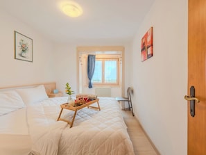 Ein Schlafzimmer mit Bett, Nachttisch, Fenster mit Vorhängen, einem Sessel und einem Tablett mit Gegenständen.