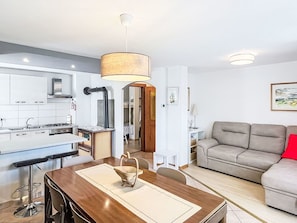 Eine moderne Küche mit weißen Schränken, ein Essbereich mit einem Holztisch und ein graues Sofa mit einem roten Kissen.