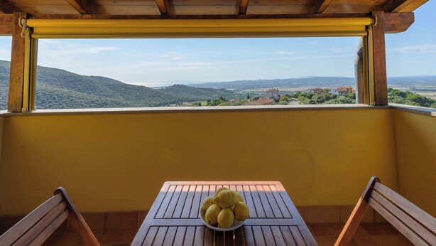 Un balcó de fusta amb una taula i cadires, un bol de limones i una vista panoràmica de turons i edificis.