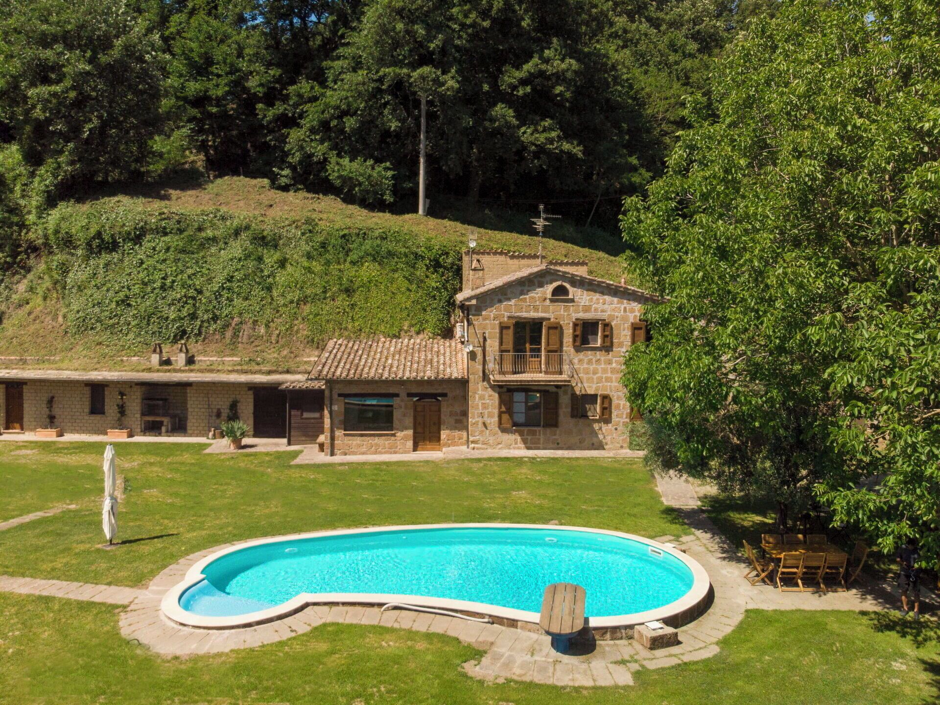 A stone building with a swimming pool, surrounded by greenery and a hilly landscape.